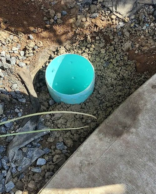 A teal relief-well standpipe cap set into a gravel bed beside a concrete walkway.