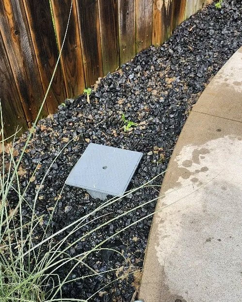 A square drain access cover set discreetly into a gravel and mulch bed along a fence line, fully landscaped and restored.