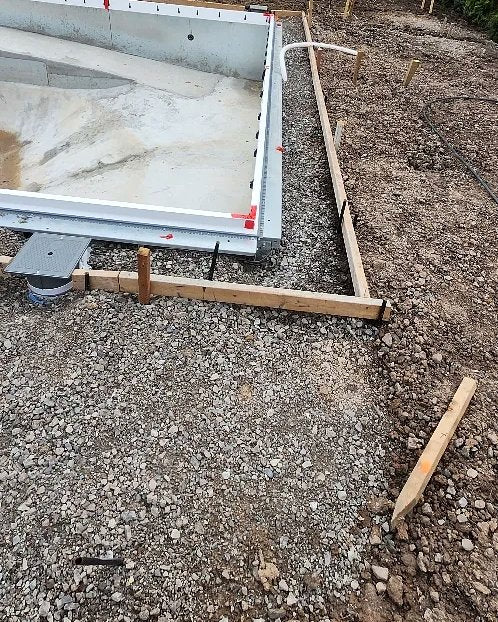 A grey relief-well access cover set in gravel at the corner of a new vinyl-liner pool shell, before the surrounding soil is backfilled.